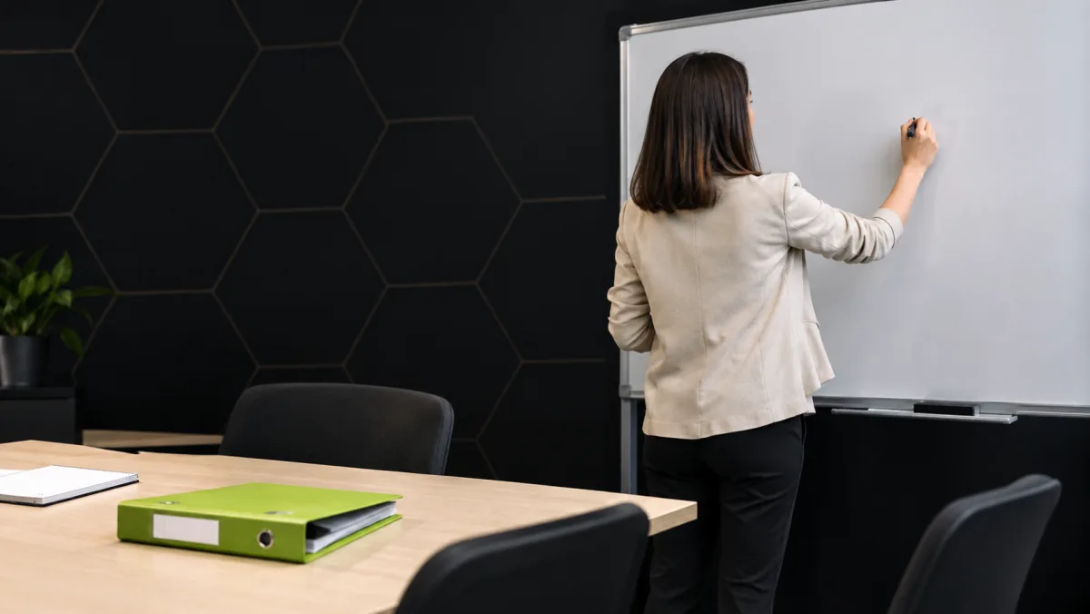 Une formatrice rédige les objectifs d'un programme de formation sur un tableau blanc, un classeur vert lime posé sur la table de travail.